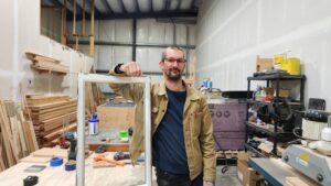 Vance Lump standing in his workshop holding a window he is working on.