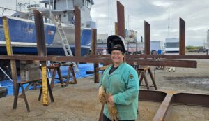 Sara Ruscetta in her welding gear standing in front of the outrigger rack she is welding