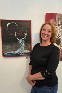 Naomi Roberts stands next to her painting of a person with antlers sitting on the ground with an eclipse in the sky