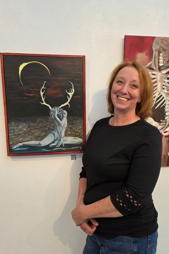 Naomi Roberts stands next to her painting of a person with antlers sitting on the ground with an eclipse in the sky